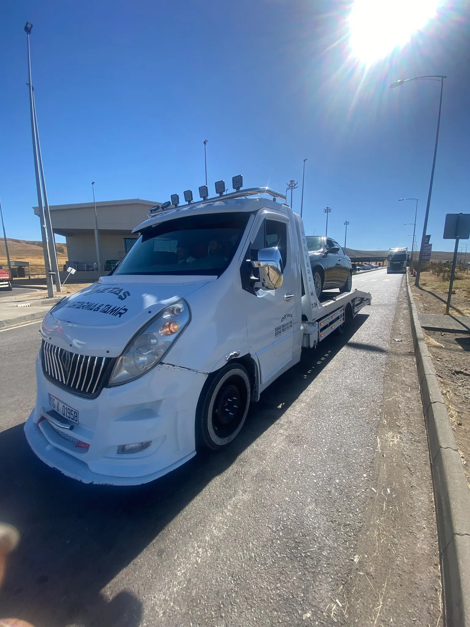 Güneşli bir günde otoyol tesisinin yanında, platformuna station wagon yüklenmiş oto çekici.