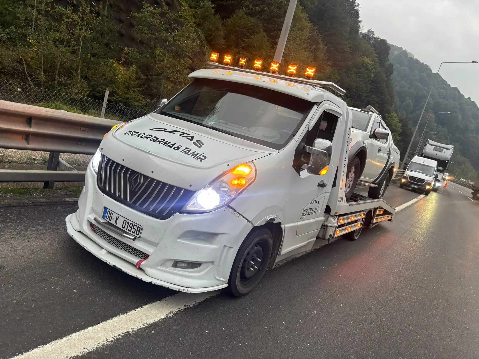 MYK Oto Servis'in kayar kasa çekicisi, sabah saatlerinde otoyolda platformunda bir pikap taşıyor; tepe lambaları yanıyor.