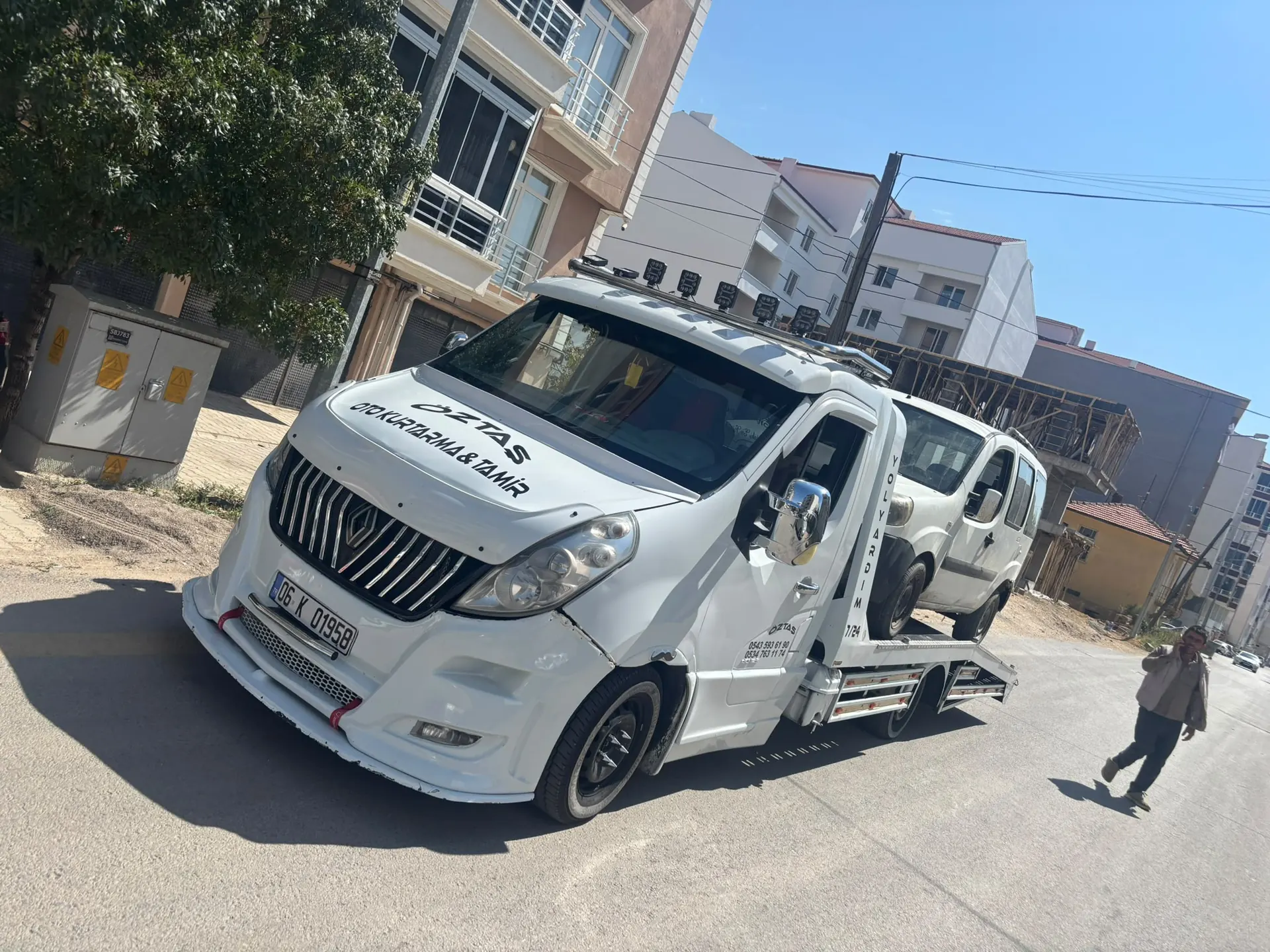 İlçe merkezinde bir sokakta, platformuna beyaz hafif ticari araç yüklenmiş oto çekici.