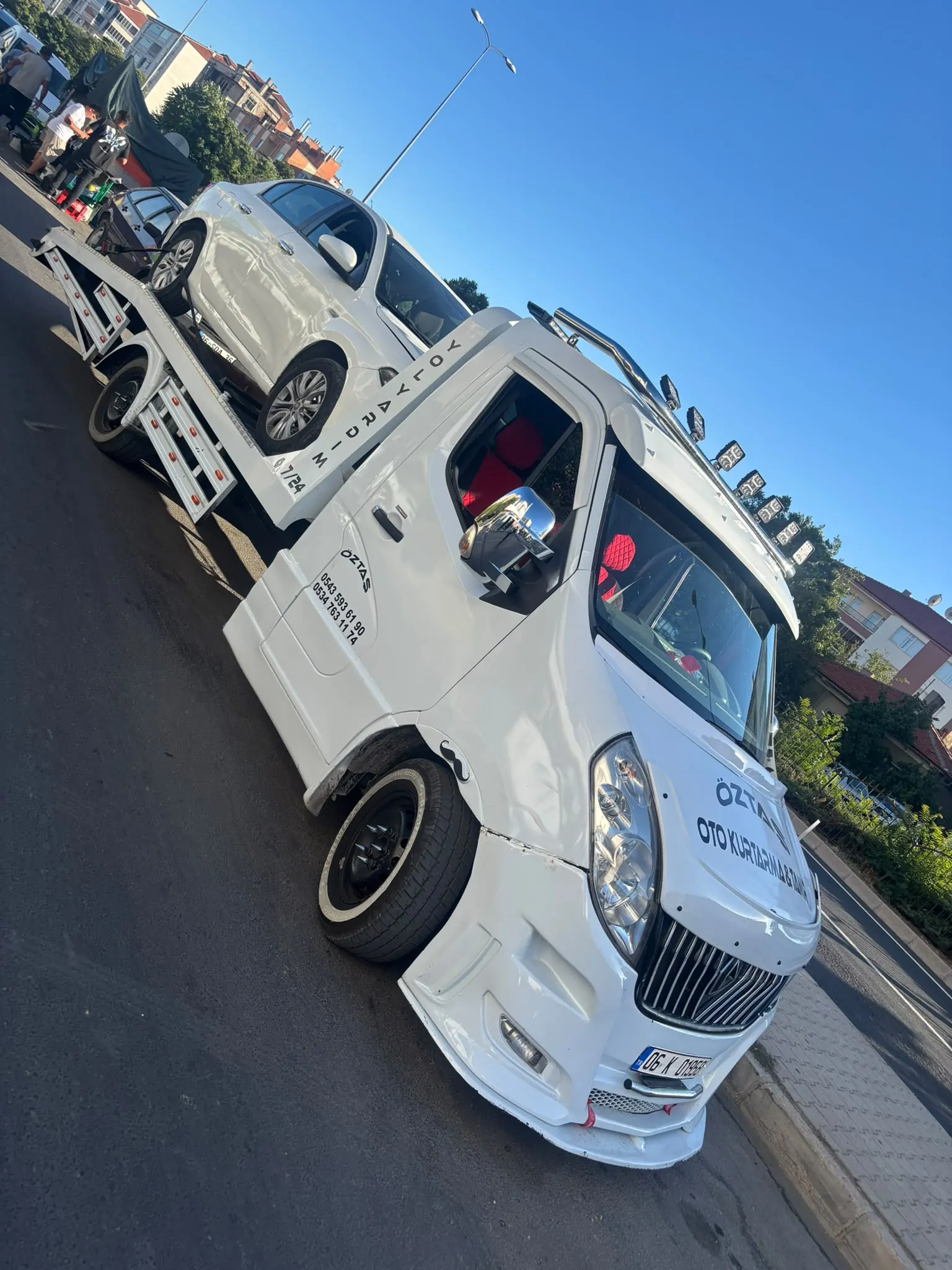Şehir içinde yol kenarında duran oto çekici, platformuna beyaz bir hatchback yüklenmiş.