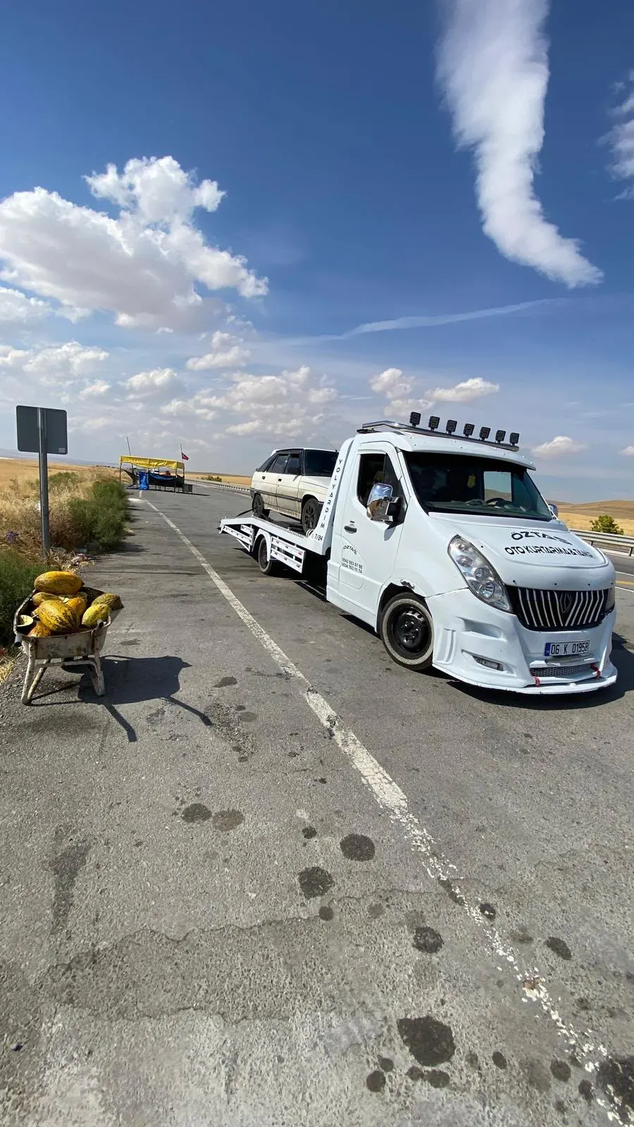 Devlet yolu kenarında, kavun tezgâhının yanında platformuna eski bir otomobil yüklenmiş oto çekici.